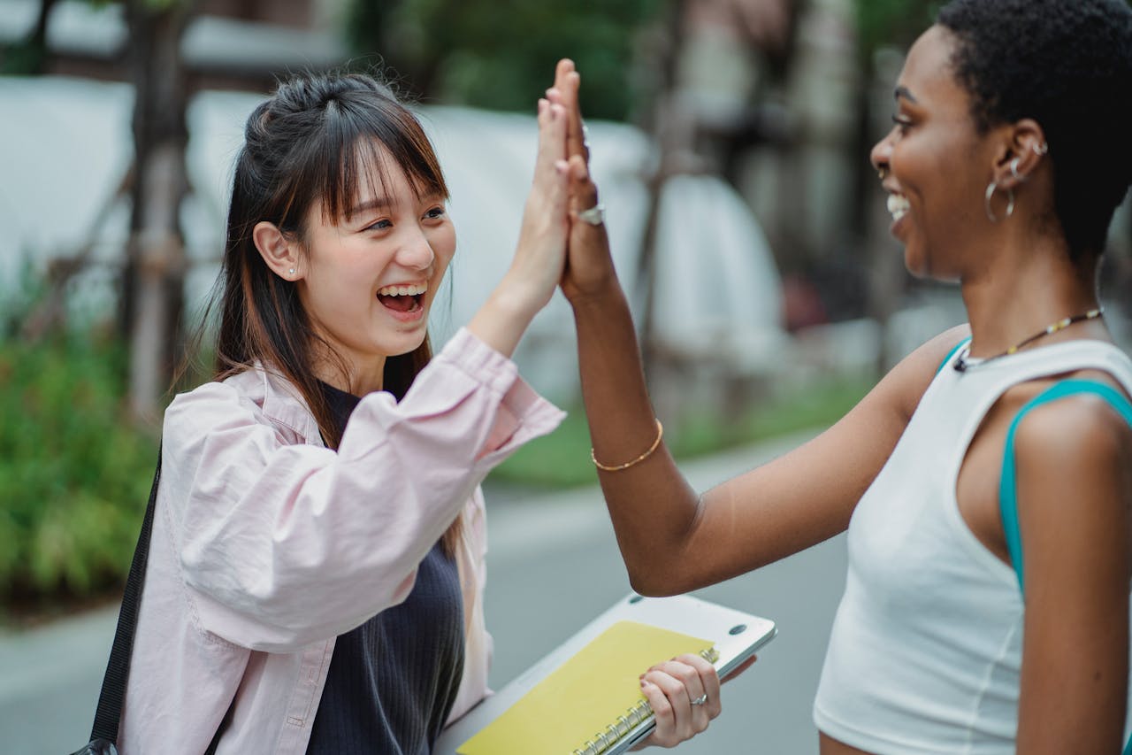 Side view excited multiracial female students in casual clothes giving high five to each other while spending break in lush park